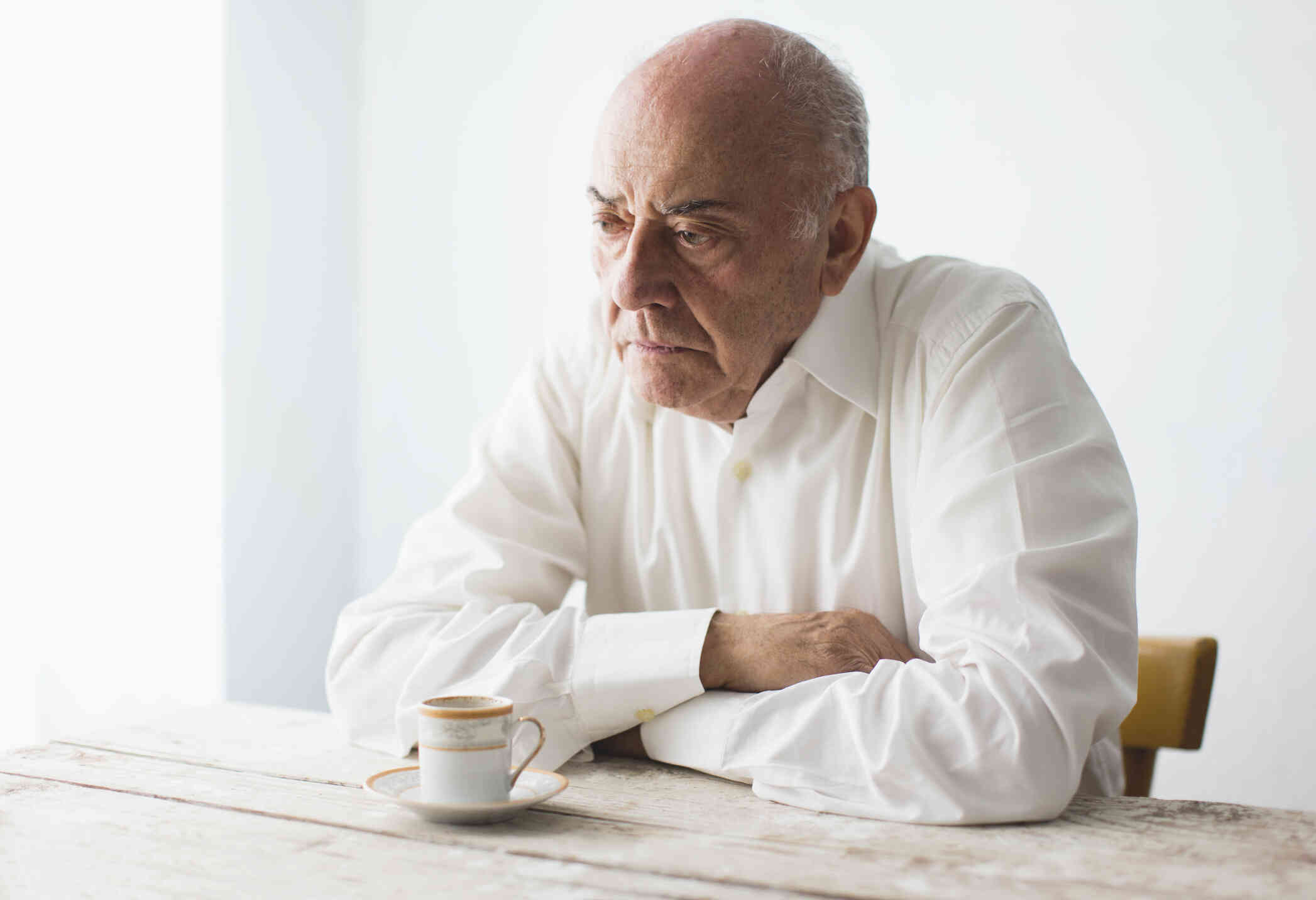 An elderly man in a white button down shirt sits hunched over at the table and crosses his arms while gazing off sadly.